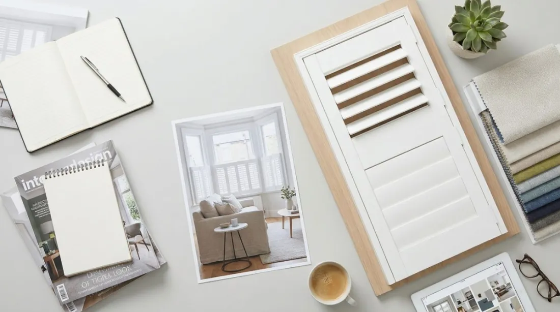 A bright living room bay window fitted with white tier-on-tier plantation shutters, lower panels closed and upper louvres tilted open, with a sofa and side table in front