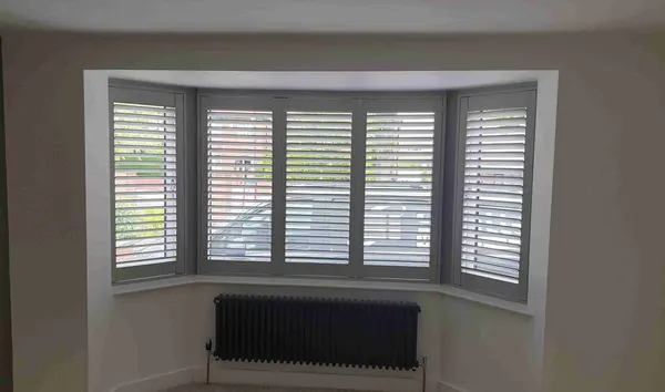 Angled bay window with white shutters above a black radiator